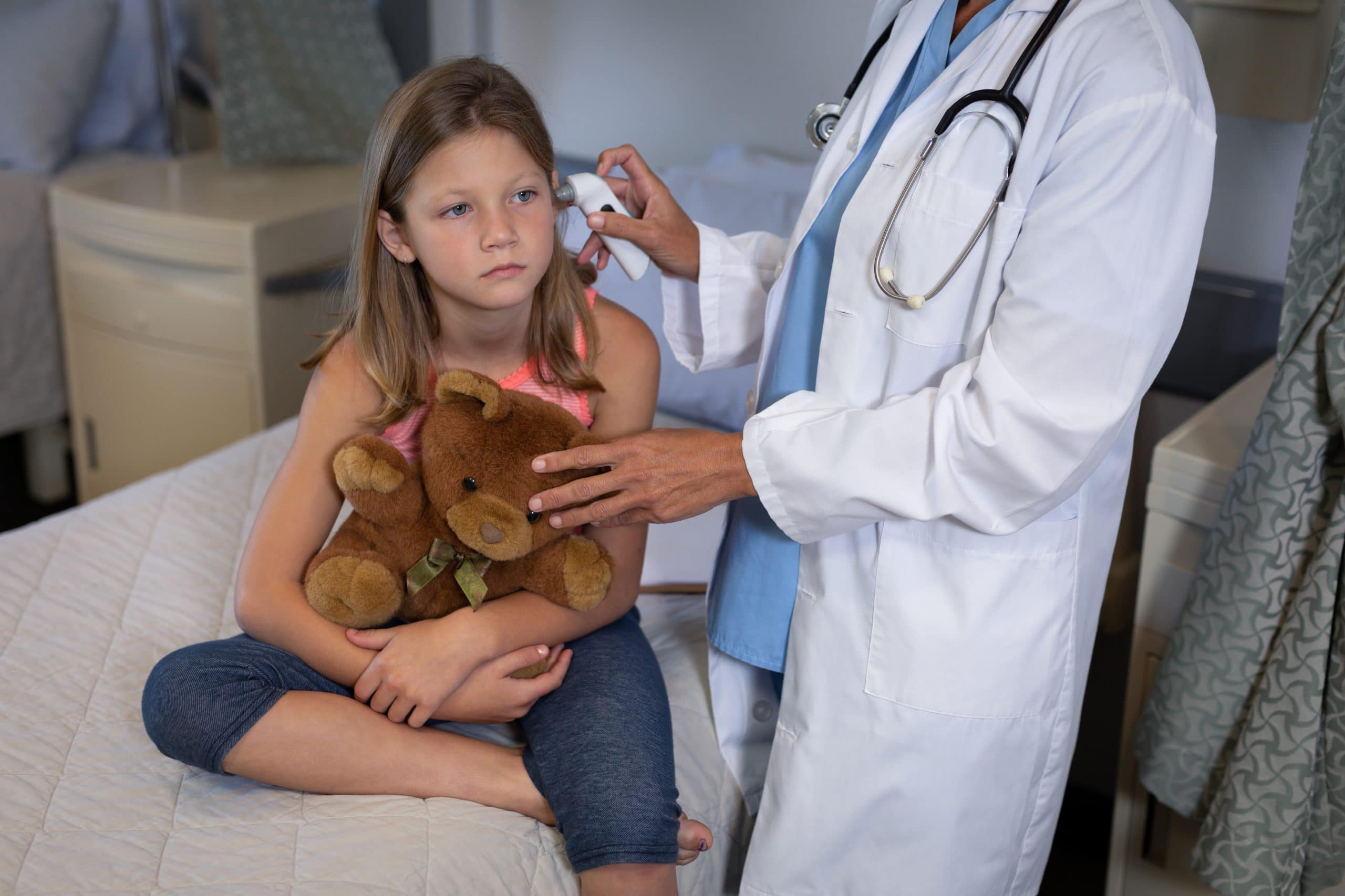 a physician in while laboratory coat with stethoscope checking her pediatric patient's temperature using an ear thermometer